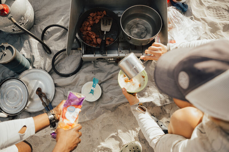 A top-down shot shows people camping and cooking outdoors. A portable stove has a pan with food and a pot of water on it. Someone is pouring something from a metal container into a bowl. Other camping supplies are scattered around on a blanket or tarp, including plates, utensils, and a propane tank.